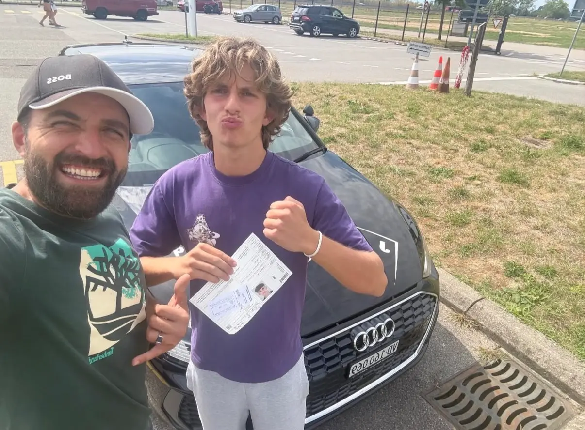 Mergim with his student in a purple T-shirt, holding their driving licence beside the Just Drive Audi.