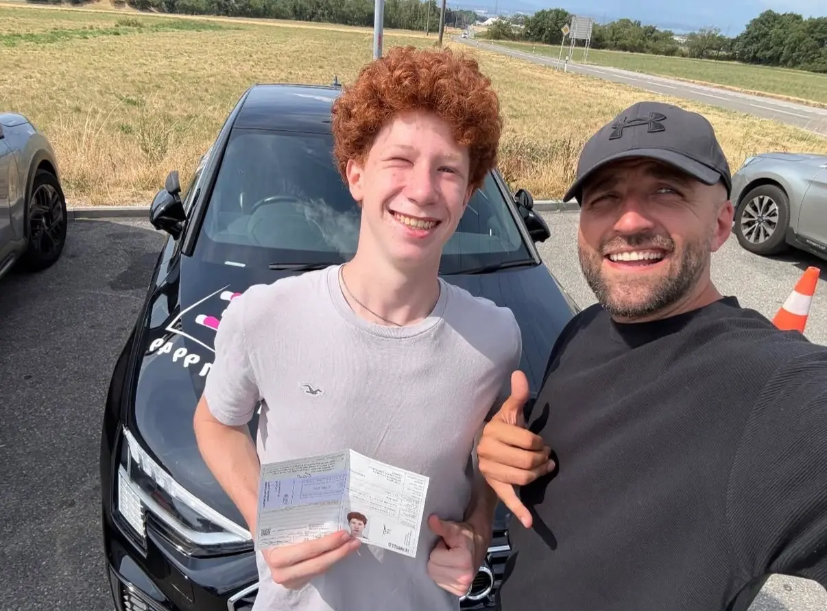 Mergim and his student in a grey T-shirt, holding a document beside the Audi.
