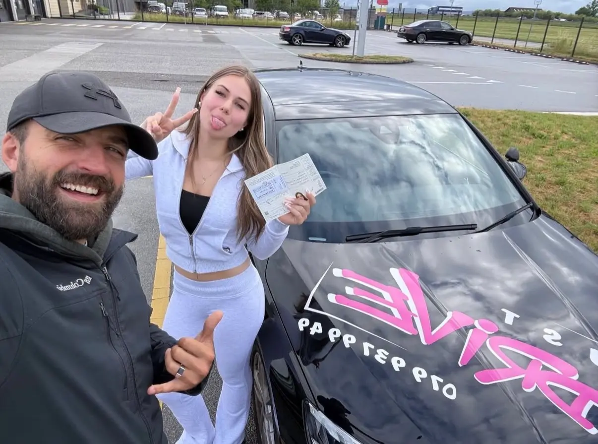 Mergim and his student dressed in white, holding their driving licence beside the Audi.